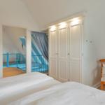 Schlafzimmer mit Bett, Kleiderschrank und Stuhl. Blick in einen Flur mit blauer Brüstung.