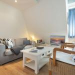 Wohnzimmer mit Sofa, zwei Stühlen und Fernseher. Holzboden und Fenster mit blauen Vorhängen.