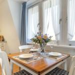 Essbereich mit Tisch, Stühlen und Blumen. Fenster mit weißen und blauen Vorhängen.
