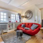 Ein Wohnzimmer mit rotem Sofa, Holzboden und Fenstern mit Vorhängen.