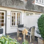 House with thatched roof, white door and windows, wooden table with chairs in the garden.