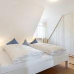 Schlafzimmer mit Doppelbett, weißen Wänden und Holzboden. An der Wand steht ein weißer Schrank.