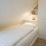 Schlafzimmer mit Einzelbett, weißen Wänden und schräger Decke. Weiße Schrankwand und Nachttisch mit Lampe.