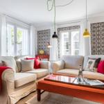 Two sofas with cushions and blanket, table with glass top, windows with white curtains and patterned walls.