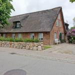Red brick house with thatched roof, white windows, stone wall, and flowering shrubs.