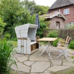 Terrace with white lounge chair, table and chairs. Background: house with reed fence and flowers.
