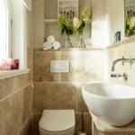 Badezimmer mit Toilette, Waschbecken und Fenster. Wandregale mit Blumen und Handtüchern.
