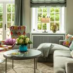 Ein Wohnzimmer mit Sofa, Sessel und Tisch. Fenster und Vorhänge sind sichtbar.