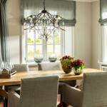Esszimmer mit Holztisch, Stühlen und Kronleuchter. Fenster mit Rollläden und Blumenarrangements.