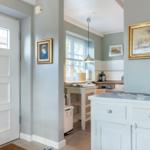 House with kitchen area and entrance door. Wall pictures and white furniture.