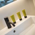Drei gelbe Natur-Duschgels, Shampoo und Lotion auf einer weißen Waschschale mit schwarzer Wasserhahn.