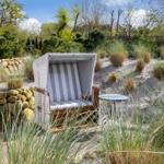 White beach chair with striped cushions on sandy ground with grasses and stone wall.