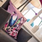 A bird-patterned cushion on a chair near a window with flowers.
