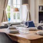 Dining room with wooden table, chairs, and table setting. Window with curtains and TV in background.