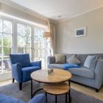 Living room with sofa, blue armchairs, and wooden table. Windows overlook the garden.