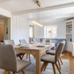 Dining room with wooden table, chairs, and windows. Bookshelf and ceiling light visible.