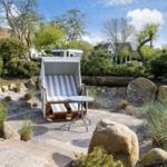 Garden with beach chair, table, and stones. Background: trees and houses.