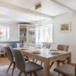 Dining room with wooden table, chairs, and windows. Bookshelf and lamp visible.