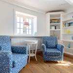 Two blue patterned armchairs are by a window. White shelves are on the wall.