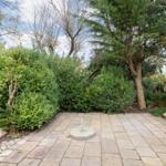 Garden with stone paving, trees, and shrubs. Central metal post on stone base.