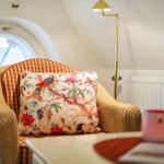 A chair with patterned upholstery and a cushion with bird and flower motifs in front of a round window.