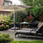 Garten mit Sonnenschirm, Tisch, Stühlen und Liegestühlen. Blumenbeete und Steinmauer umgeben den Platz.
