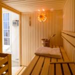 Sauna mit Holzbänken, Holzfußboden und Holzwand. Dampfheizung und Holzkrug mit Löffel.