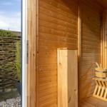Holzsauna mit Einstiegsbereich und Außentür. Holzbank und Eimer stehen bereit.