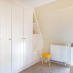 Schlafzimmer mit weißem Schrank, gelbem Hocker und Fenster.