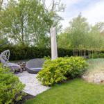 Garten mit Sitzbereich, Grasfläche und Sträuchern. Hintergrund: Bäume und Hecke.
