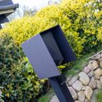 Schwarzer Briefkasten im Garten neben Sträuchern und Steinmauer.