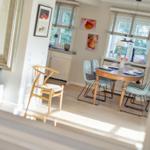 Dining room with wooden table and chairs, windows with blinds, wall art, and living area in background.