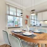 Dining room with wooden table, chairs, and windows. Kitchen with white cabinets.
