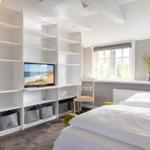 Schlafzimmer mit Bett, TV und Fenster. Weiße Regale und grauer Teppich.