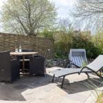 Terrasse mit Tisch, Stühlen und Liegen. Hintergrund: Holzzaun und Grünflächen.