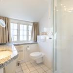 Bad mit Dusche, Waschbecken und Toilette. Fenster mit Vorhängen.