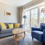 Wohnzimmer mit grauem Sofa, blauem Sessel und Holztisch. Fenster mit Blick auf Außenbereich.
