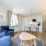 Wohnzimmer mit Essbereich und Küche. Blaue Sessel, Holztisch, Fenster mit Vorhängen.