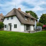 House with thatched roof, terrace, and garden. Wooden tables and chairs outdoors.