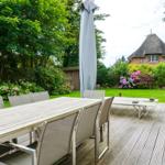 Wooden terrace with table, chairs, and umbrella. A house with thatched roof in the background.