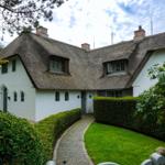 A white house with thatched roof, green lawn, and paved path.