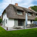House with thatched roof, terrace, and garden. Table, chairs, and umbrella are outside.
