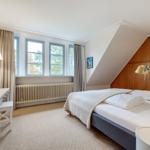 Schlafzimmer mit Bett, Schreibtisch und Fenstern. Holzschrank an der Wand.