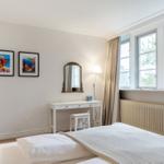 Schlafzimmer mit Bett, Schreibtisch und Fenstern mit Blick auf Bäume.