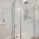 Dusche mit Glaswand, Duschbrause und Handbrause. Wand mit Haken und weißer Tasche.