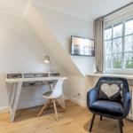 A bedroom with a desk, chair, and armchair by a window.