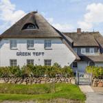 Weißes Haus mit Strohdach und Gartenzaun. Schriftzug GREEN TEFT auf Fassade.