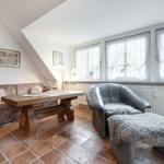 Wohnzimmer mit Sofa, Holztisch und Ledersessel. Terrakottaboden und Fenster mit Vorhängen.
