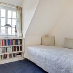 A bedroom with sloped ceiling, bed, bookshelf, and window with curtains.