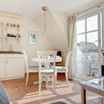 Dining room with white table, chairs, and kitchen area. Large windows with view to balcony.